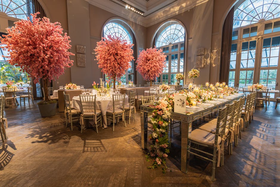 Elegant indoor setting featuring large pink cherry blossom trees in substantial dark planters positioned behind round tables and a long rectangular table, all decorated with a variety of fresh floral 
