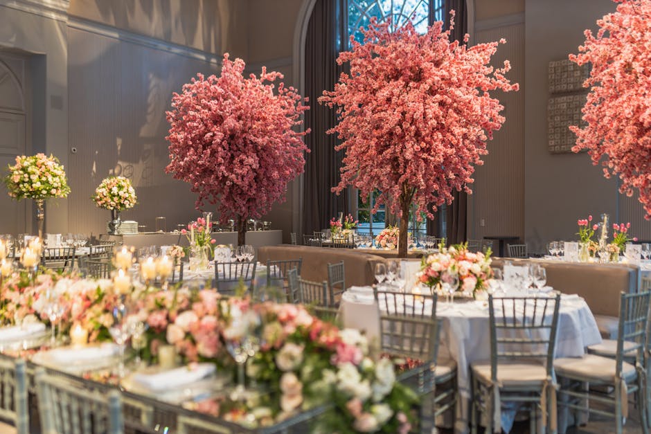 An elegant event space decorated with large, vibrant pink cherry blossom trees serving as focal points, surrounded by round banquet tables draped with white tablecloths and adorned with lush floral ar
