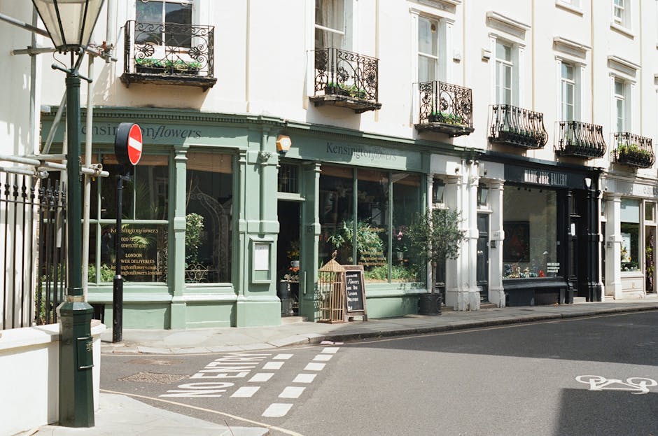 A floral shop named Kensington Flowers situated on a street corner in Fitzrovia. The shop's exterior is painted in pale green with large glass windows displaying various flower arrangements inside. Po